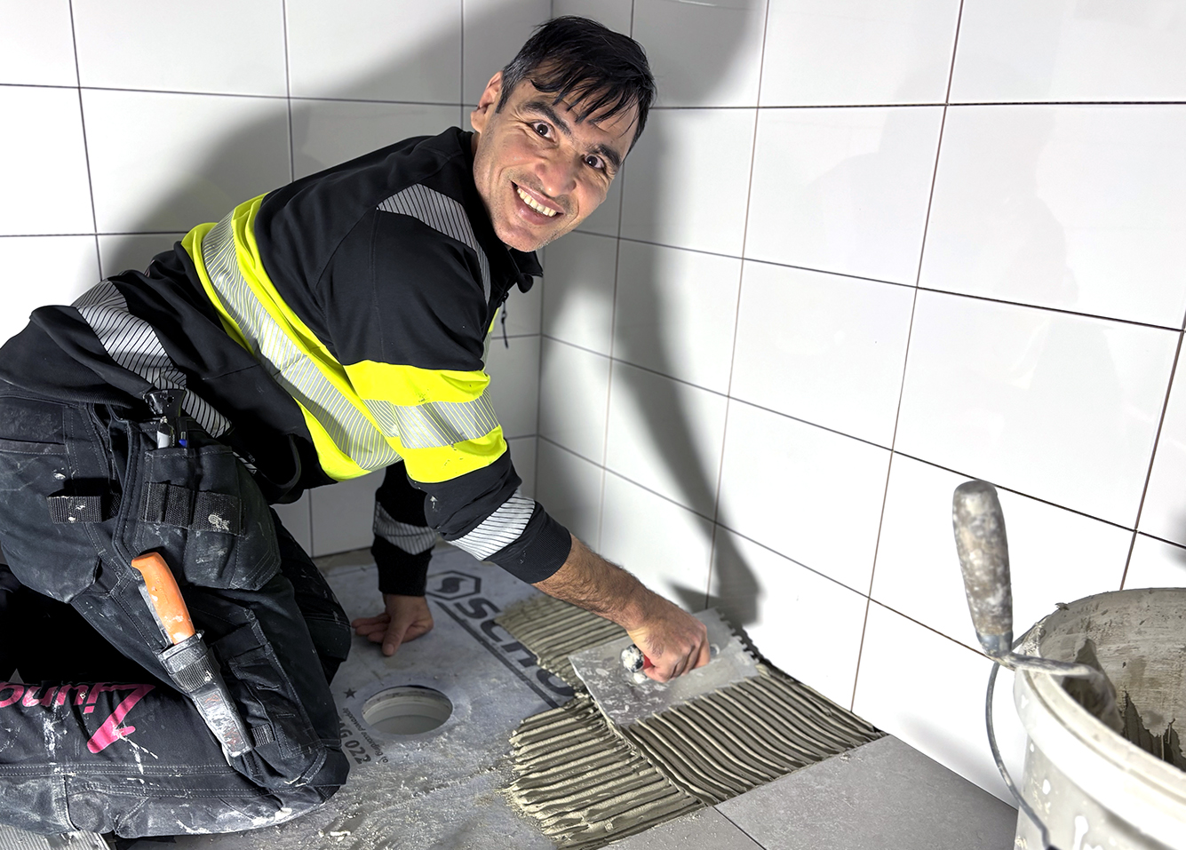 Man arbetar med plattsättning i badrum och tittar leende in i kameran