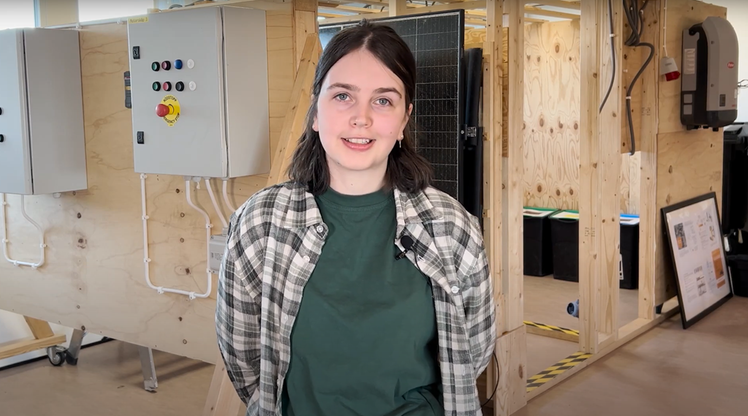 Ung tjej i rutig skjorta med grön tshirt under står i verkstadsmiljö.