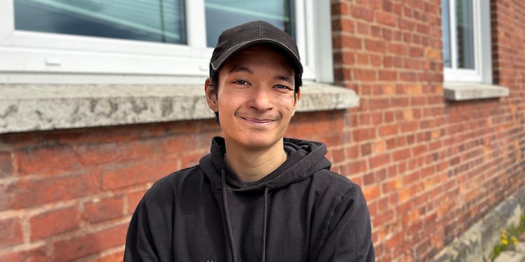 Ung kille står i svart hoodie och keps framför ett tegelhus.