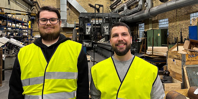 Två män i reflexvästar står i en produktionslokal.